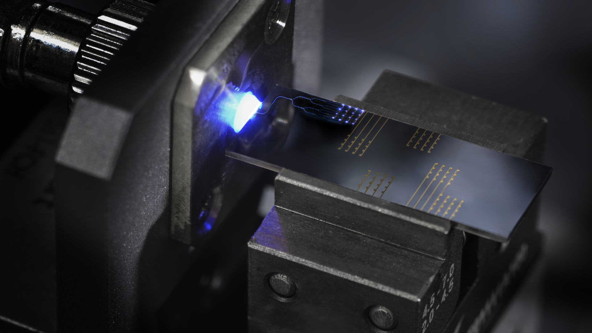 A close-up shot shows a small, rectangular circuit board clamped securely in a mechanical fixture. A bright blue light illuminates the edge where a delicate ribbon cable connects to the surface of the board, which features intricate gold-colored traces.