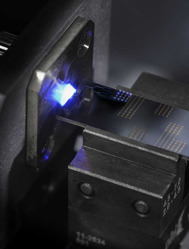 A close-up shot shows a small, rectangular circuit board clamped securely in a mechanical fixture. A bright blue light illuminates the edge where a delicate ribbon cable connects to the surface of the board, which features intricate gold-colored traces.