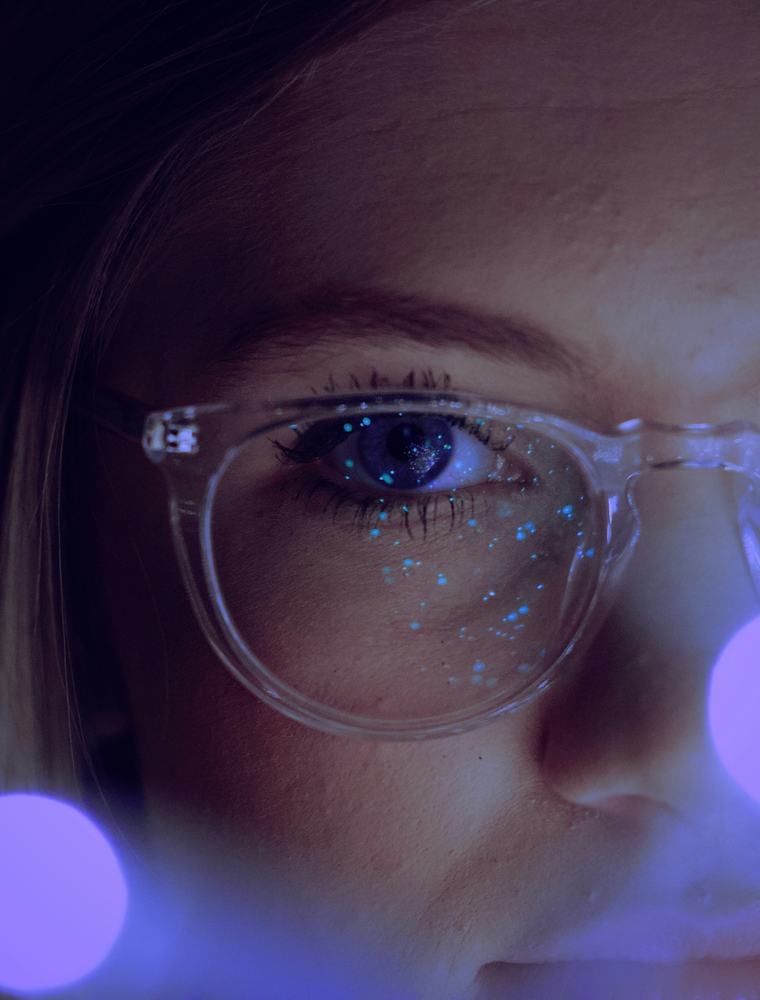 A close-up shot of a person wearing clear-framed glasses, illuminated by glowing blue light reflections.