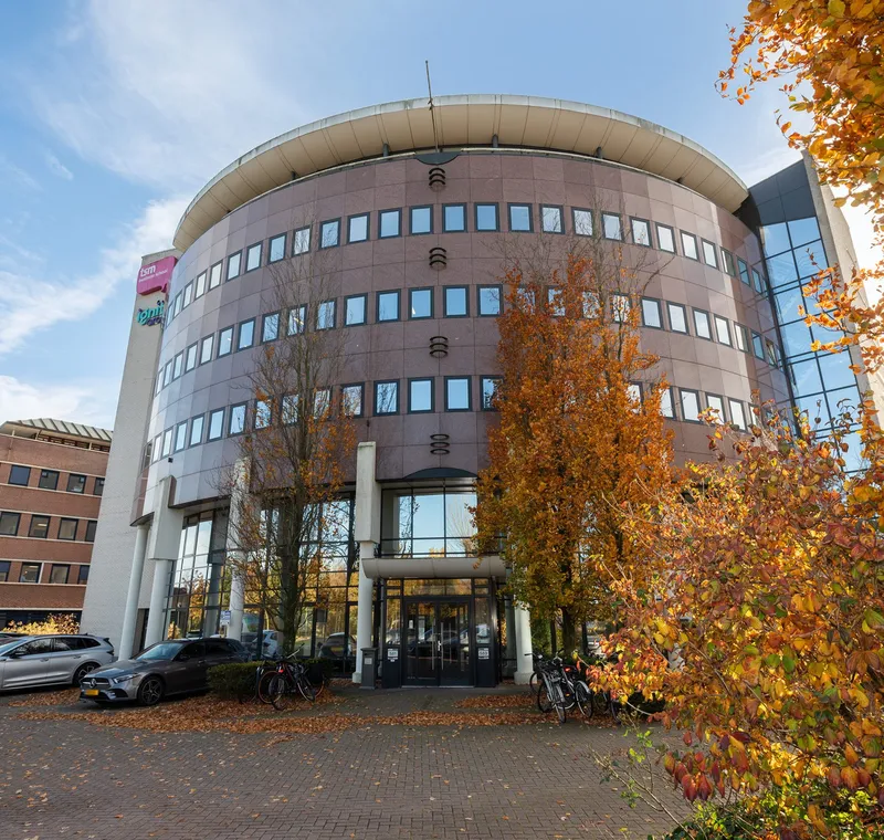 A multistory curved building features rows of rectangular windows and a circular top section, with trees displaying autumn foliage in the foreground. Parked cars and bicycles are visible near the entrance, under a partly cloudy sky.