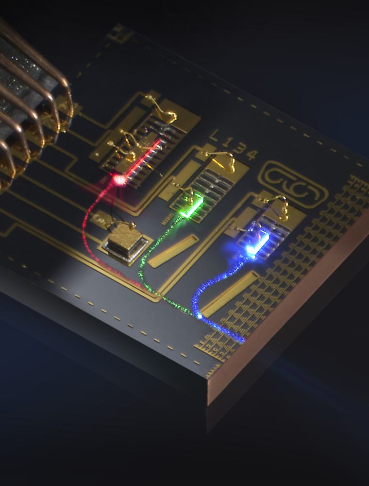 A close-up shot shows a photonic chip with glowing red, green, and blue light paths. A bundle of copper wires connects to the side of the circuit board.