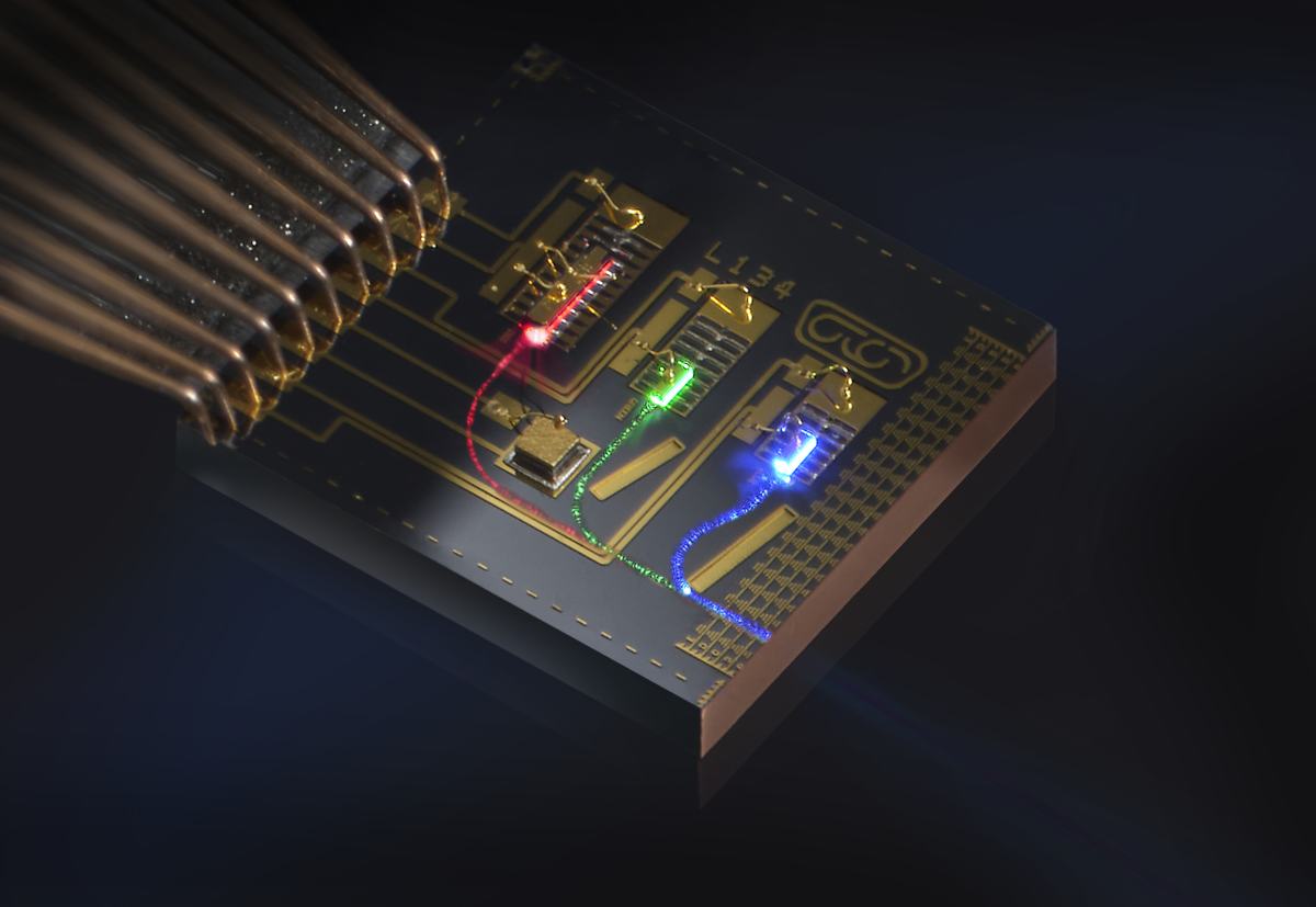 A close-up shot shows a photonic chip with glowing red, green, and blue light paths. A bundle of copper wires connects to the side of the circuit board.