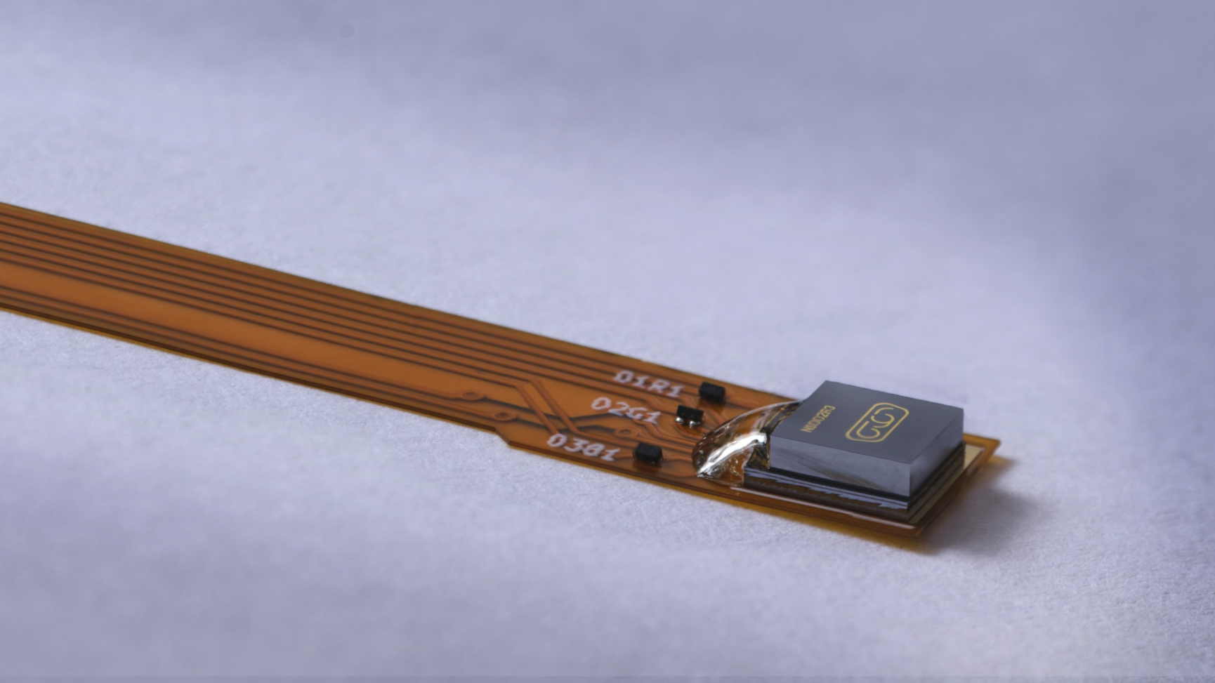 A long, thin circuit board rests on a grey surface. The brown strip has text printed near its end and several parallel circuit traces running its length. A small component is mounted near the opposite end.
