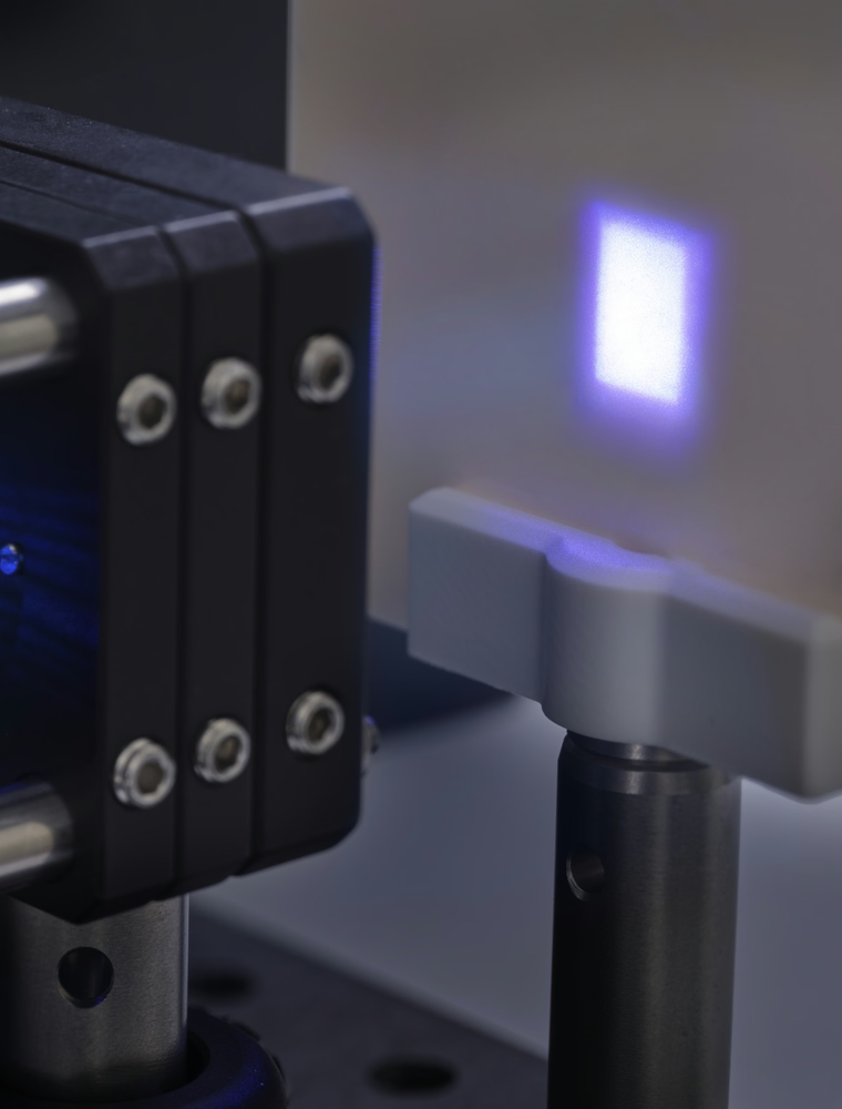 A medium shot of an optical setup, focusing on a Thorlabs mount holding a lens or sensor, with blue light passing through it and projecting onto a white surface.