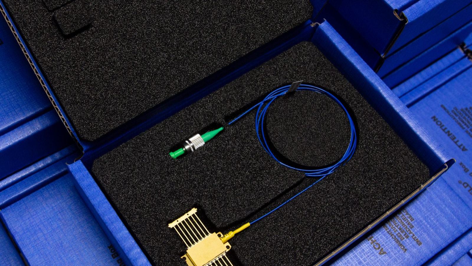 An open blue box lined with black foam rests on a table, holding a gold-colored electronic component connected to a blue fiber optic cable with a green connector and a coiled section.
