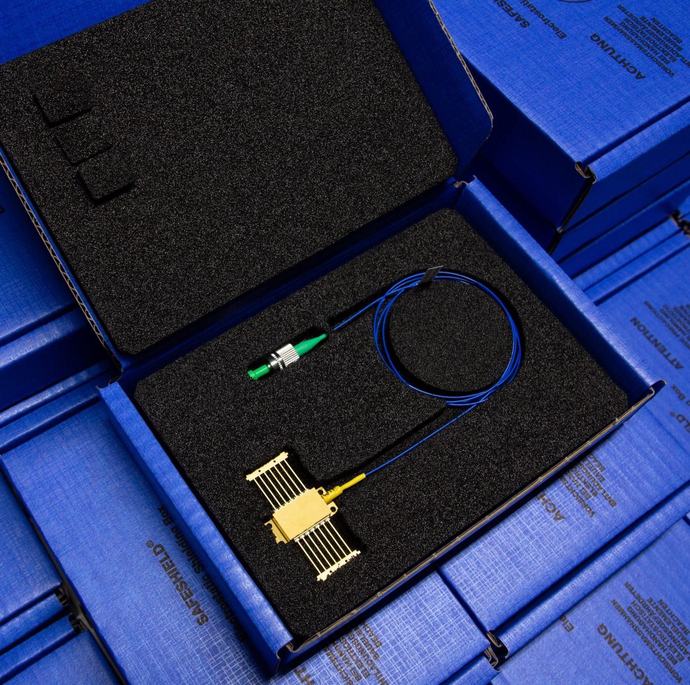 An open blue box lined with black foam rests on a table, holding a gold-colored electronic component connected to a blue fiber optic cable with a green connector and a coiled section.