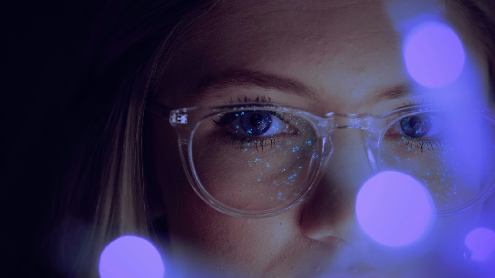 A close-up of a person wearing clear-rimmed glasses, with glowing blue lights reflecting across the frames and face, creating an out-of-focus bokeh effect in the foreground.