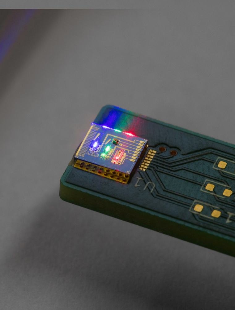 A small electronic circuit board features an illuminated microchip on one end, with several gold-colored contact pads labeled D1, D2, and D3. The chip glows with vibrant blue, green, and red lights, casting a colorful reflection onto a nearby smooth…