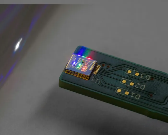 An eye-level shot captures a small circuit board resting on a white surface, featuring a green base and a component with blue and green lights.