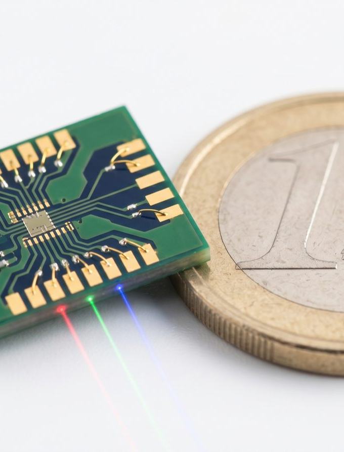 A green circuit board sits next to a coin for scale, with red, green, and blue light beams extending from one edge.