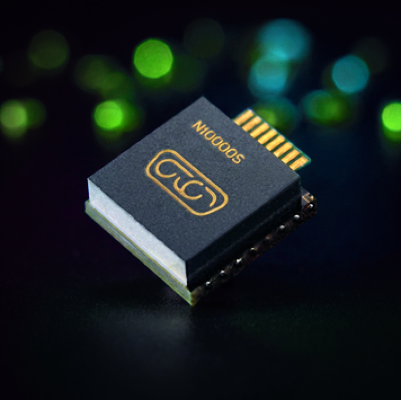 A close-up shot of a small electronic component featuring a dark top surface with gold-colored symbols and text. The component is angled against a dark background, with soft, blurred green and blue lights scattered in the background.