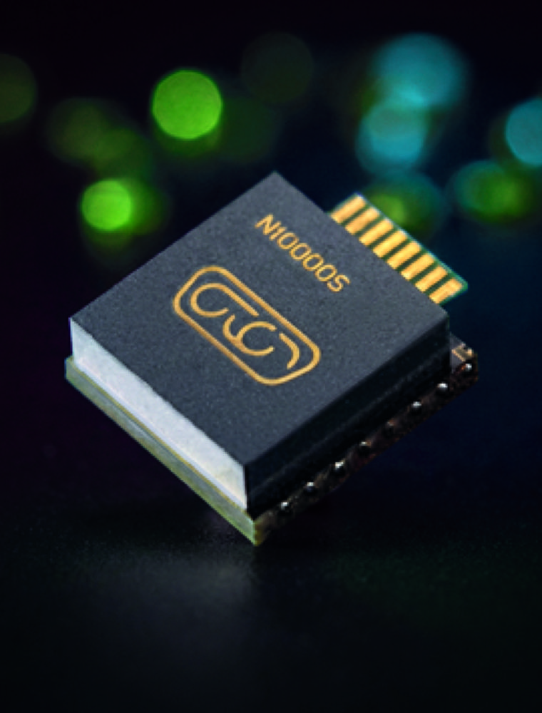 A close-up shot of a small electronic component featuring a dark top surface with gold-colored symbols and text. The component is angled against a dark background, with soft, blurred green and blue lights scattered in the background.