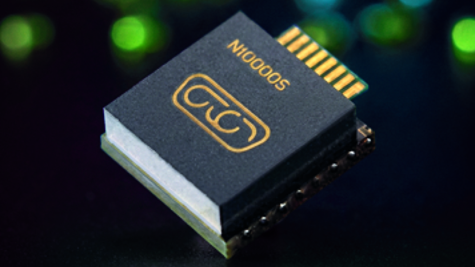 A close-up shot of a small electronic component featuring a dark top surface with gold-colored symbols and text. The component is angled against a dark background, with soft, blurred green and blue lights scattered in the background.