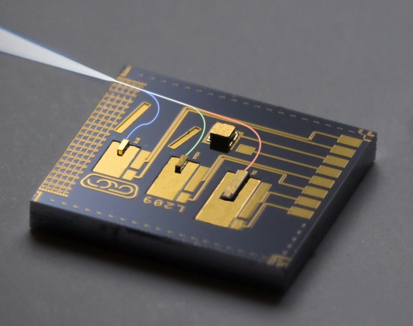 A rectangular circuit board rests on a dark surface, featuring gold-plated metallic traces and components. A thin tool points toward the board, near curved blue and red light paths.