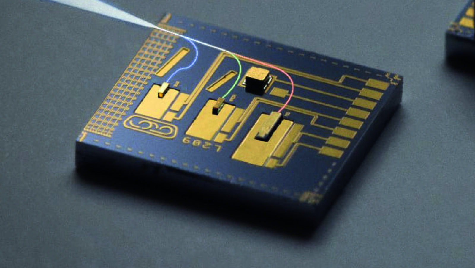 A close-up shot shows a microchip with gold traces and components on a dark background. A pointed instrument enters from the upper left, touching the chip with a thin, glowing fiber that traces lines across the surface.